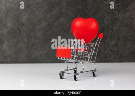 Red heart in shopping cart on a white table on dark grey background. Stock Photo