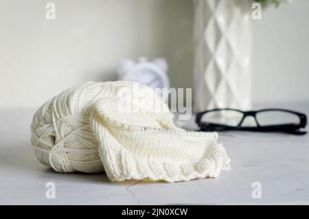 Light beige knitting wool, knitting needles and eyeglesses lay on grey table . Female hobby knitting. Soft woolen yarns for knitting warm clothes Stock Photo