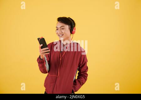 Studio portrait of cheerful young Asian man wearing headphones when watching video on smartphone Stock Photo