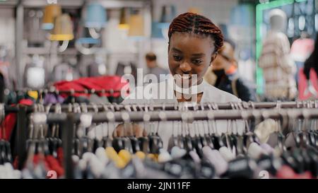 Happy rich smiling african american girl woman customer buyer chooses in store trending fashionable luxury clothes making choice looks on prices Stock Photo