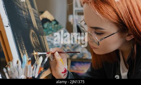 Side view red-haired painter artist creator designer woman student girl in glasses holding brush paints picture on black background with golden oil Stock Photo