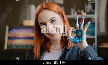 Red-haired girl talented artist female painter stands posing in art studio workshop looking at camera show okay gesture dirty palm in acrylic oil Stock Photo
