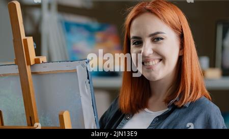 Caucasian woman artist girl painter with red hair enjoying art work with paints creates picture drawing painting on canvas looking at camera smiling Stock Photo