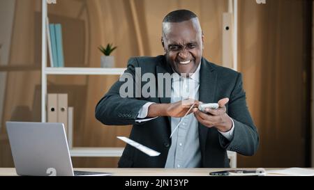 Happy successful old African American businessman senior mature man worker investor in office sitting at table holding fan of money throwing dollars Stock Photo