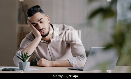 Tired sleepy hispanic man arab guy dreaming overworked male student slumber at office laying down on desk leisure close eyes napping feel exhausted Stock Photo