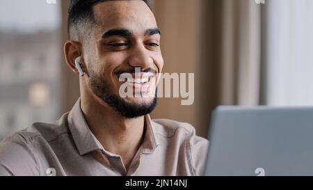 Joyful young arabic man wear wireless earphones look at laptop relax watch funny video comedy film smiling enjoy free break time read good news Stock Photo