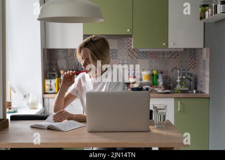 Serious middle-aged woman sits in kitchen making notes about job vacancies in paper pad Stock Photo