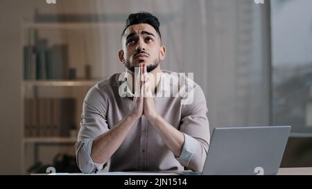 Frustrated young hispanic male analyst religious business man feel stress check financial documents ask for help concentrate mind at problem sit with Stock Photo