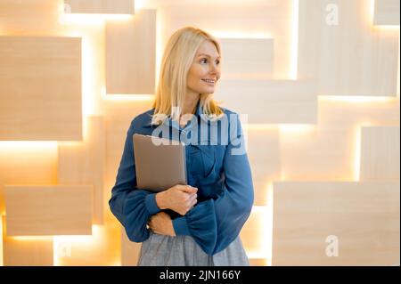 Successful intelligent elegant mature Caucasian woman, business lady, in business clothes, female secretary, holding a laptop in her hand, standing in the modern office, looking to the side, smiling Stock Photo