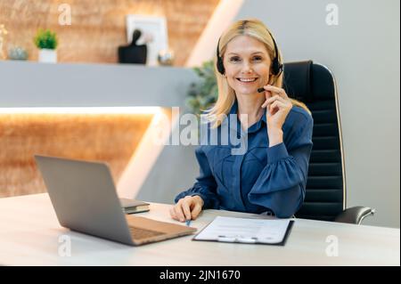 Portrait of elegant intelligent mature caucasian businesswoman, female call center operator, financial advisor, in formal wear, with headset, sits in modern office, looks at camera, smiling friendly Stock Photo