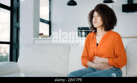 Sad African woman at home curly hair ill girl sitting on sofa in kitchen living room holding belly touching abdomen suffering from stomachache painful Stock Photo