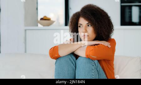 Alone lonely stressed African American woman sitting with sad problem at home couch thinking about trouble looking away bad health stress frustrated Stock Photo