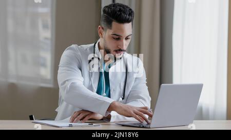 Concentrated arabian man doctor practitioner sit at clinic cabinet use computer chatting distant in medical social app give online consultation Stock Photo