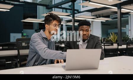 Two multicultural professional male co-workers men colleagues sit at office use laptop discuss internet project hold negotiation choosing business Stock Photo