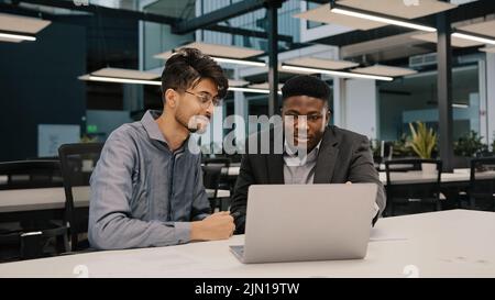 Two multicultural professional male co-workers men colleagues sit at office use laptop discuss internet project hold negotiation choosing business Stock Photo