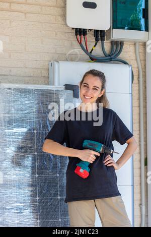 Vertical view of young happy smiling hard working woman looking to camera installing the solar panel system with batteries and a electric screwdrivers Stock Photo