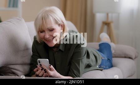 Old happy Caucasian grandmother 60 years senior woman 50s lady blonde elderly female lies on couch at home enjoys remote mobile conversation video Stock Photo