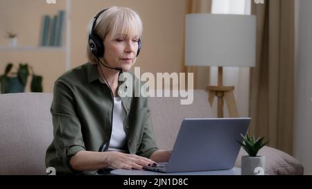 Middle aged business woman in headset talking video conference distant meeting remote calling with laptop. Senior old Caucasians female online teacher Stock Photo