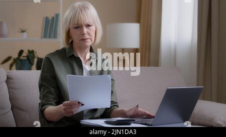 Caucasian old 50s homeowner housewife makes woman budget checks monthly income by checking paper notes using app laptop. Middle-aged businesswoman Stock Photo