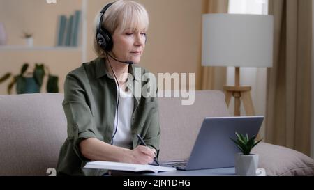 Senior businesswoman in headphones sitting on couch listen online training webinar distant e-learning 60s woman business class video chat call Stock Photo