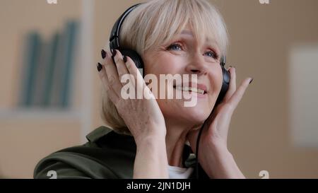 Portrait happy relaxed blonde 60s grandmother dancing old Caucasian woman in headphones carefree senior lady in earphones listening to music enjoys Stock Photo