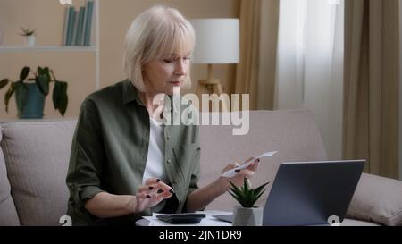 Serious caucasian old senior woman sitting on sofa checking financial calculation money budget bill pay with online app managing bank finances calcula Stock Photo