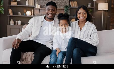 African family sit down on comfortable couch in living room. Biracial parents and child smiling looking at camera. Cheerful daughter with mum and dad Stock Photo