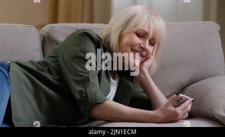 Happy relaxed Caucasian old senior mature woman holding smart phone looking at cellphone laughing lying on sofa couch enjoying using mobile apps for Stock Photo