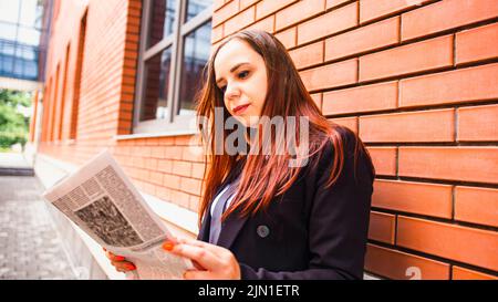 Young woman reading newspaper on street. Side view of young woman with long hair in casual clothes standing on street and reading newspaper. Stock Photo