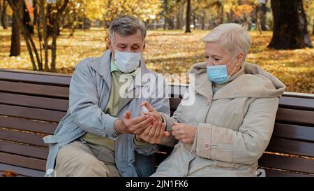 Elderly couple in medical masks in autumn park spouses disinfect hands with antiseptic sitting on bench retirees care about health personal hygiene Stock Photo