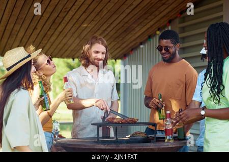 Portrait of bearded young man grilling meat outdoors with diverse group of friends at barbeque party in Summer Stock Photo