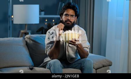 Serious focused Arabic Hispanic Indian bearded man guy wearing glasses 30s male with popcorn watching TV at home sofa late evening night relaxing Stock Photo
