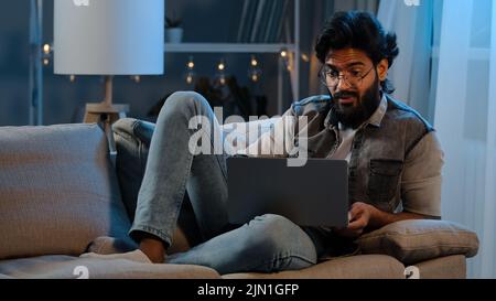 Businessman guy Arab Indian man bearded male user wearing glasses sitting on sofa at night late time with laptop reading bad news online trouble app Stock Photo