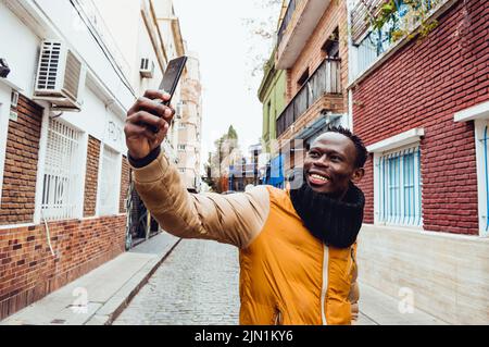 young man of african ethnicity standing in the middle of the street smiling taking a selfie with his mobile phone. Stock Photo