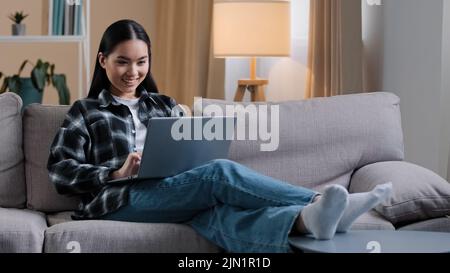 Smiling asian woman using laptop casual browsing searching online play computer game. Korean student girl freelancer sitting on couch e-learning Stock Photo