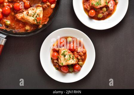 Cod stew with chickpeas, cherry tomatoes and olives over dark stone background. Top view, flat lay Stock Photo