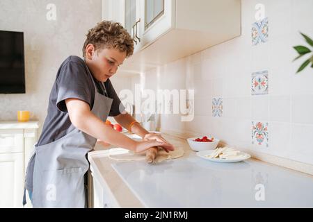 An 11-year-old boy rolls pizza dough in the kitchen. Stock Photo