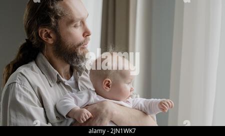 Close-up caucasian father bearded daddy holding baby daughter son infant newborn looking out window with curtains dad blowing to short blonde hair of Stock Photo