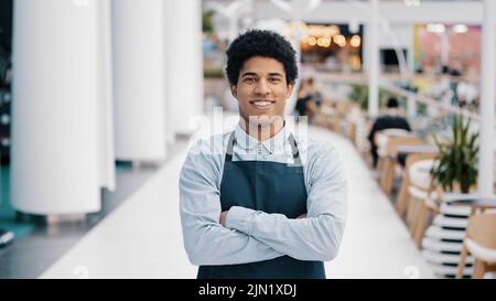 Confident African 30s restaurant worker biracial guy strong position standing indoors barman seller waiter in apron looking to camera successful cafe Stock Photo