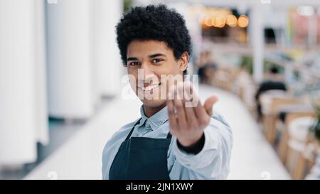 Friendly african american young guy male waiter salesman restaurant cafe kitchen bar worker in apron looking at camera showing gesture her you come Stock Photo