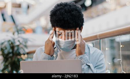 African american sad business man guy student wearing medical mask working with laptop in corporate office holding head feeling headache suffering Stock Photo