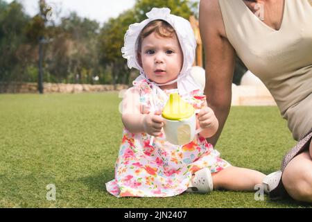 A cute little baby girl toddler wearing a white bonnet and a dress sat on green grass with her grandmother and holding a sippy cup with water Stock Photo