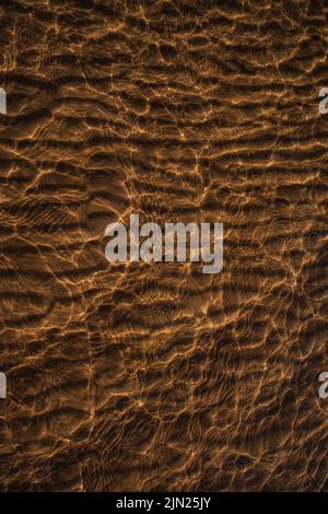 Clear sea water with golden brown sand beach and sundown sunlight that reflects abstract texture on water surface. Stock Photo