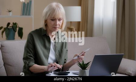 Caucasian old sad homeowner housewife makes woman budget checks monthly income by checking paper check using app laptop. Middle-aged businesswoman Stock Photo