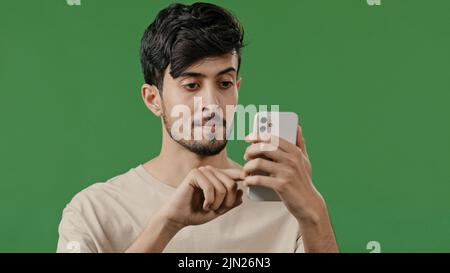 Portrait close-up arabian young man in studio on green background holding phone happy smiling guy browsing social networks make online order read news Stock Photo