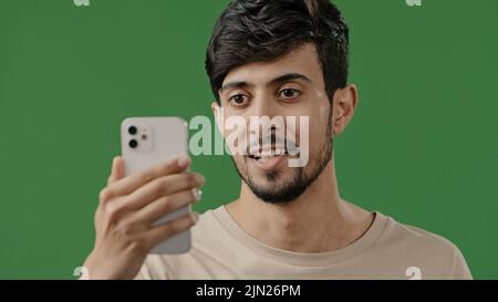 Arabic young smiling guy making video call on green background in studio using conference app on phone joyfully talking with friend online shares good Stock Photo