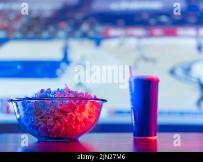 A sports match on the screen of a large plasma TV. Popcorn in a glass bowl and a drink in a plastic cup. Classic sports fan fast food set. Advertising Stock Photo