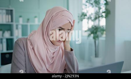 Arabian worker user Islamic businesswoman Muslim in hijab in office sitting table looking laptop screen reading bad news failure lost job mail sad Stock Photo