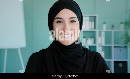 Female portrait happy Arabian 20s girl in black hijab Islamic Muslim woman posing indoors looking at camera looking at camera carefree friendly face Stock Photo