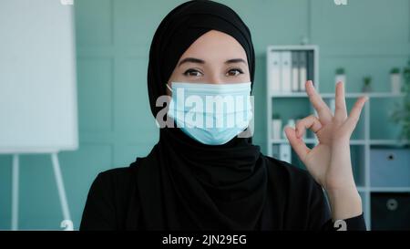 Female portrait in office Islamic successful businesswoman in black hijab wears medical mask Arab Muslim lady boss leader worker shows ok gesture Stock Photo